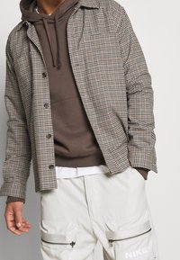 Brun rutiga jacka med knappdesign, bärs över en brun hoodie. Ljust färgade cargobyxor med dragkedjedetaljer, med Nike-logotypen.