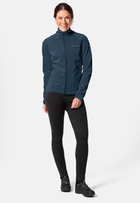 Giacca softshell blu navy con colletto alto, leggings sportivi neri e scarpe da escursionismo nere. Presenta una zip frontale e un design aderente.