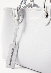 Biała torba na ramię z teksturowanej skóry z srebrnymi elementami, z zawieszką wytłoczoną napisem "Emporio Armani" oraz ozdobnym plecionym paskiem ze skóry.