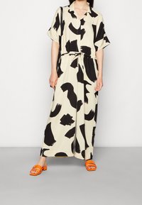 Vestido de estampado negro y crema con mangas cortas y un lazo en la cintura, combinado con sandalias naranjas de tipo deslizante. Textura de tela suave.
