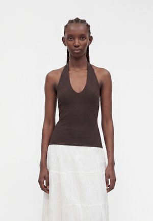Femme aux cheveux tressés portant un haut sans manches marron foncé et une longue jupe blanche à volants, debout devant un fond uni.