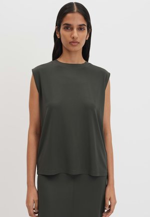Femme aux longs cheveux foncés porte un haut sans manches vert foncé et une jupe assortie, debout devant un fond clair uni.