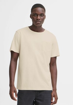 T-shirt beige à manches courtes avec un col rond et une petite poche poitrine, en tissu lisse. Porté avec un jean foncé.