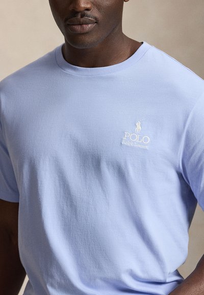 Άνδρας με γαλάζιο t-shirt Polo Ralph Lauren με λογότυπο στο αριστερό στήθος, στέκεται μπροστά σε ουδέτερο φόντο.