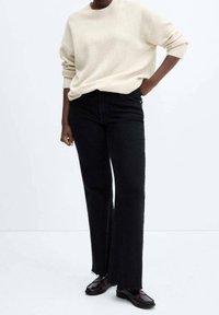 Pull en tricot crème avec un col rond, associé à un jean noir taille haute à jambes larges et des chaussures marron à lacets. Textures lisses, coupe décontractée.