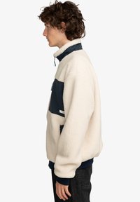 Fleece sweatshirt i creme med marineblå accenter. Har en høj krave, forlommes og ribbede manchetter. Blød tekstur, afslappet pasform.