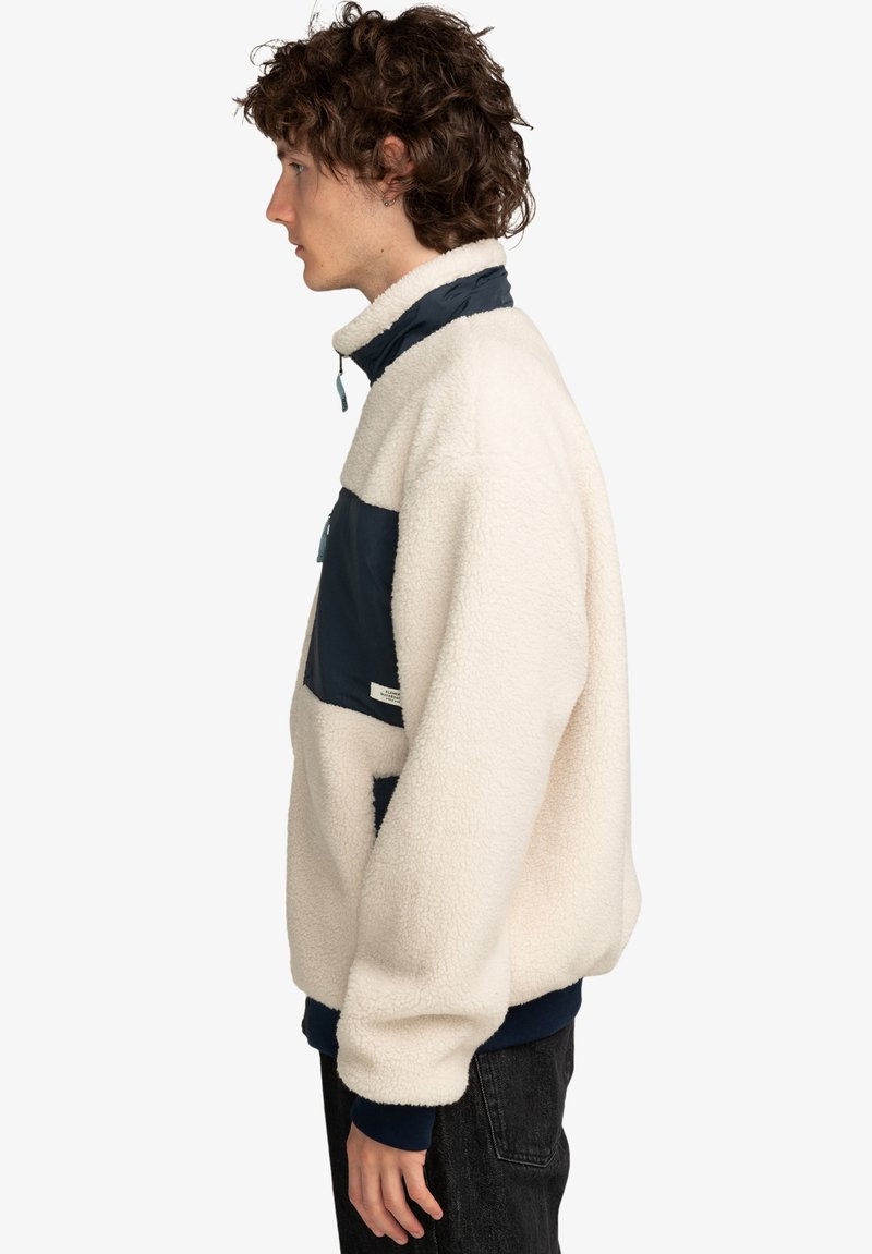 Pulóver de forro polar en color crema con acentos en azul marino. Presenta un cuello alto, bolsillo frontal y puños acanalados. Textura suave, corte relajado.