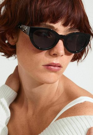 Black cat-eye sunglasses with dark lenses and studded accents on the arms, set against a light background. Texture of fabric is visible.