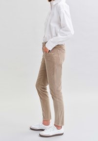 Woman wearing beige slim-fit pants, white long-sleeve shirt, and white sneakers, standing with one hand in pocket against plain background.