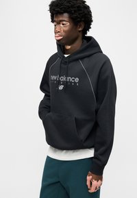 Zwarte hooded sweater met een voorzak, wit logo en grijze accenten. De stof lijkt zacht, met een ontspannen pasvorm en geribbelde manchetten.