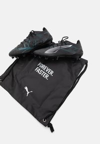 Botas de fútbol negras con diseño texturizado, acentos turquesa y logo blanco, acompañadas de una bolsa negra con cordón que presenta texto en blanco.