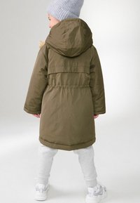 Parka căptușit de culoare verde măsliniu, cu glugă, talie elastică și mâneci lungi. Asortat cu pantaloni de trening gri și sneakers albi. Căciulă din tricot texturat.
