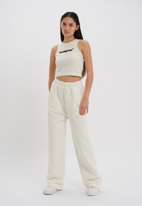 Witte cropped tanktop met zwart logo en bijpassende losse sweatpants. Glad textuur, elastische tailleband en twee zijzakken. Witte sneakers.