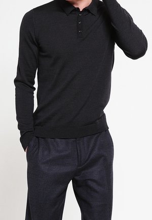 Mand iført en tætsiddende sort poloshirt med lange ærmer og mørkegrå bukser, med den ene hånd i lommen og den anden hævet nær hovedet.