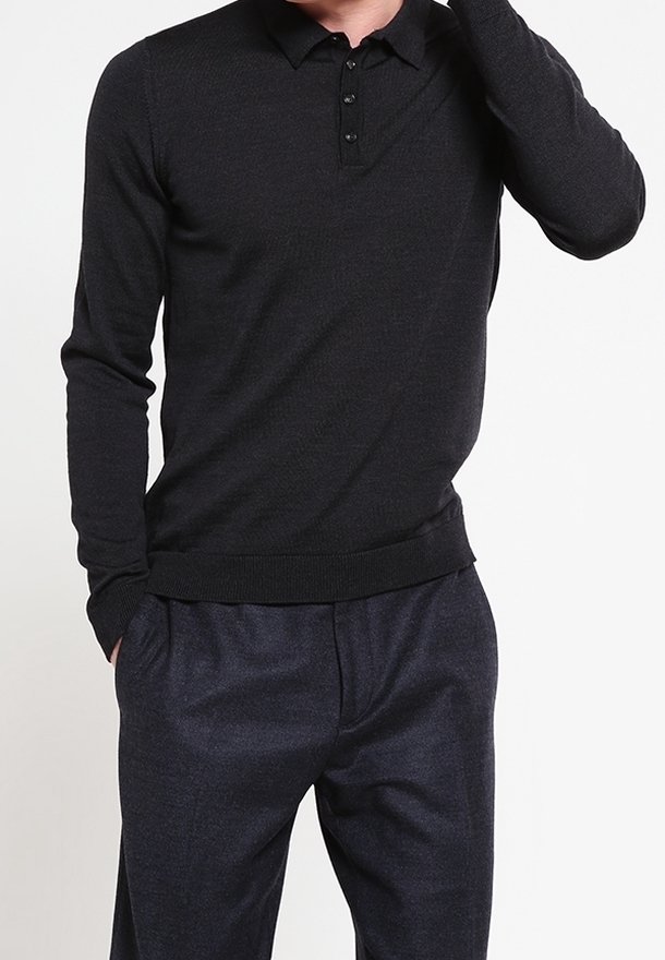 Homme portant un polo noir moulant à manches longues et un pantalon gris foncé, une main dans la poche et l’autre levée près de sa tête.