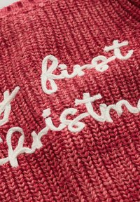 Tissu tricoté rouge avec un motif texturé et un texte brodé blanc portant la mention "premier Noël". Matériau doux et épais avec des points de couture visibles.