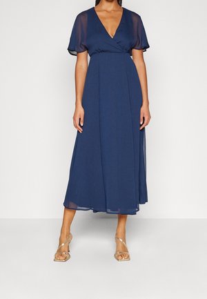 Femme portant une robe midi bleu marine avec des manches courtes en tulle transparent et un col en V, assortie à des sandales à talons hauts dorées à brides.
