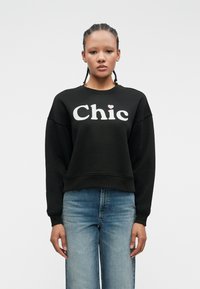 Young person with braided hair wearing black sweatshirt with "Chic" text and light blue jeans, standing against a plain white background.