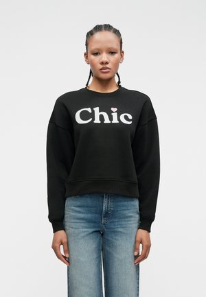 Jeune personne avec des cheveux tressés portant un sweat à capuche noir avec le texte "Chic" et un jean bleu clair, se tenant devant un fond blanc uni.