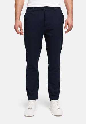 Uomo che indossa pantaloni slim fit blu navy e sneakers bianche in piedi davanti a uno sfondo neutro.