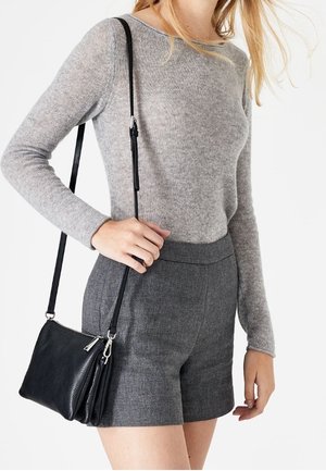 Kvinde iført en lysegrå langærmet top og mørkegrå skræddersyede shorts, der holder en lille sort læder crossover taske med lynlås.