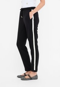 Mujer que lleva pantalones negros con franjas laterales blancas y bailarinas negras con dobles tiras y detalles de strass.