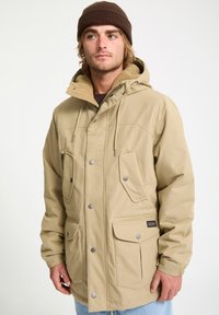 Beige hooded jacket with a soft, fleece-lined collar, multiple buttoned pockets, and silver hardware accents; smooth fabric texture.