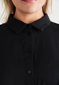 Black collared shirt with buttons, featuring a smooth texture and a fitted design. Close-up view highlighting the collar and button details.