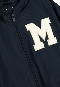 Giacca della Marina con zip frontale e cappuccio. Presenta un grande logo "M" color crema sul petto; il tessuto sembra liscio e leggero.