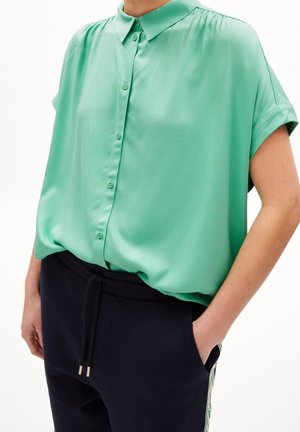 Osoba ubrana w miętowozieloną, krótkorękawową bluzkę na guziki, wpuszczoną do czarnych spodni, z rękami w kieszeniach.