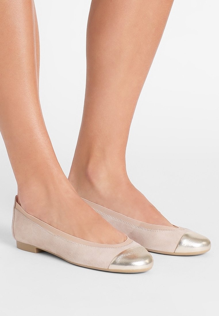 Pieds portant des ballerines en daim beige avec embouts métalliques dorés sur fond blanc.