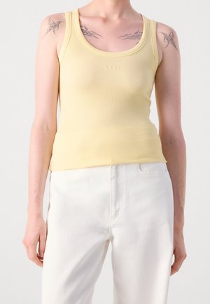 Personne portant un débardeur côtelé jaune pâle et un pantalon blanc, montrant des tatouages aux épaules et une position détendue du bras sur un fond uni.