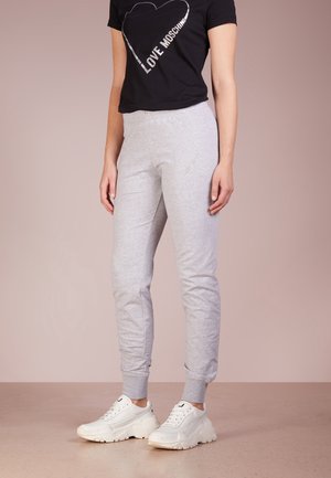 Frau trägt ein schwarzes T-Shirt mit Herz und Schriftzug, hellgraue Jogginghose und weiße klobige Sneakers, steht auf einem beigefarbenen Boden.