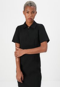 Black dress with a short collar, short sleeves, and a front zip closure. Features a fitted silhouette and side pockets. Smooth texture.