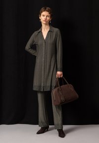 Femme portant une longue chemise à motifs et un pantalon assorti tient un grand sac à main marron, debout devant un fond noir.