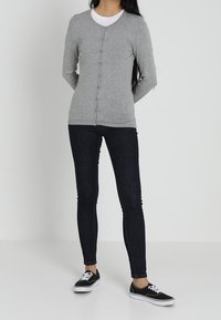 Žena stojí s rukama za zády, na sobě má šedý propínací svetr, bílou košili, tmavé skinny džíny a černé tenisky.