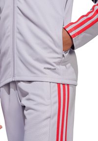 Hellgraue Sportjacke mit roten Streifen an den Ärmeln, Reißverschluss und einem "AEROREADY"-Label nahe dem Saum. Das Material wirkt glatt und atmungsaktiv.