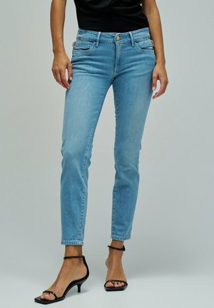 Mujer con vaqueros ajustados azul claro y sandalias negras de tacón con tiras, de pie frente a un fondo neutro.