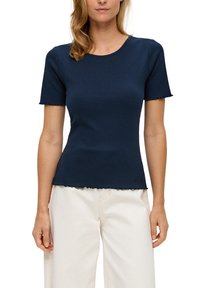 Vrouw in een aansluitend navyblauw T-shirt met een korte mouw en gegolfde randen, gecombineerd met hooggetailleerde off-white broek op een witte achtergrond.