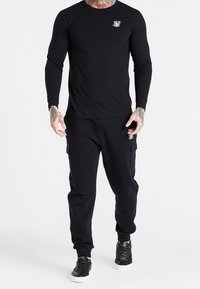 Mustapaita, jossa on pitkät hihat ja pieni valkoinen logo rinnassa, yhdistettynä mustiin cargo-housuihin, joissa on sivutaskut, sekä mustiin lenkkareihin.