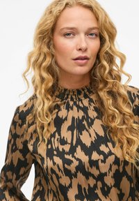 Blusa a maniche lunghe in un design a stampa animale beige e nera, con un colletto alto arricciato e un tessuto morbido e strutturato.