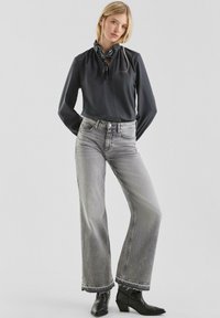 Schwarze Bluse mit langen Ärmeln und gerafftem Kragen, kombiniert mit grauen Jeans mit hoher Taille und weitem Bein. Schwarze Stiefeletten mit spitzen Zehen.