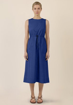Frau, die nach vorne blickend steht und ein ärmelloses, königsblaues Midikleid mit Kordelzug in der Taille sowie braune, flache Riemchensandalen trägt.