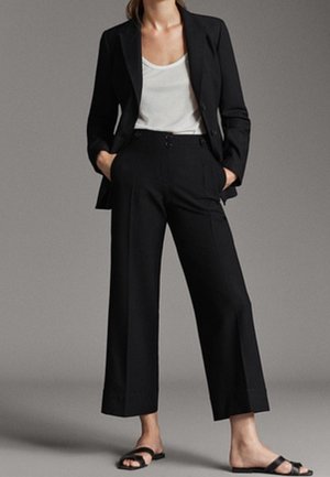 Femme portant un blazer noir et un pantalon noir à jambes larges avec un top blanc et des sandales plates noires à bout ouvert, mains dans les poches.