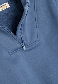 Sudadera de forro polar de color azul claro con media cremallera, textura suave y cuello alto. Presenta un tirador de cremallera y detalles de costura a lo largo de las costuras.
