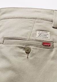 Beżowe bawełniane spodnie z tylną kieszenią, z brązowym guzikiem, białą metką z czarnym tekstem oraz czerwonym logo Levi's. Gładka tekstura.