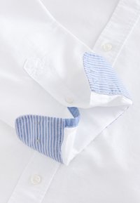 Weißer Hemdblusenkragen und Manschetten mit blau-weiß gestreiftem Innenfutter, die Knopfdetails und Stoffstruktur zeigen.