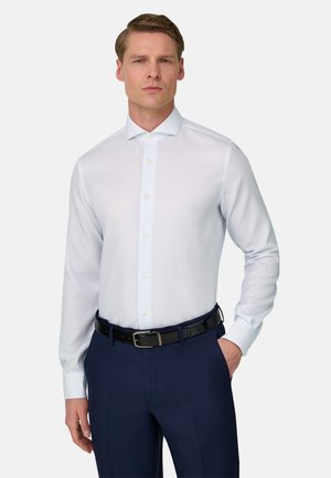 Mężczyzna ubrany w białą koszulę, granatowe spodnie i czarny pasek, stojący z jedną ręką w kieszeni na jednolitym jasnym tle.