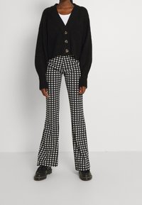 Woman wearing black cropped buttoned cardigan, white top, black and white houndstooth flared pants, and black loafers, standing against white wall.