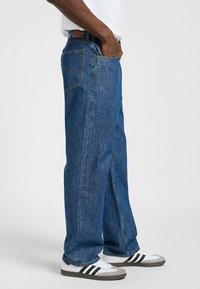 Jeans de mezclilla azul holgados con una cintura alta, cinco bolsillos y costuras amarillas. Combinados con zapatillas blancas con rayas negras.